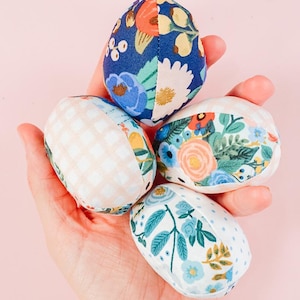 May include: Four fabric Easter eggs in a hand. The eggs are decorated with different floral patterns, including a blue and white floral pattern, a pink and white gingham pattern, a white and green floral pattern, and a white and orange floral pattern.