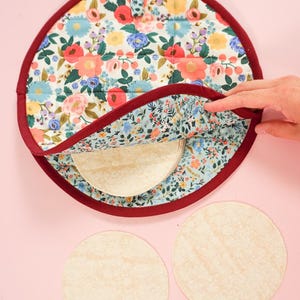 May include: A floral patterned tortilla warmer with a red trim. The warmer is open and shows a stack of tortillas inside. Two additional tortillas are on a pink surface.