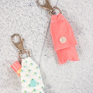 Pode incluir: Dois chaveiros de tecido com fechos de metal. Um chaveiro é rosa e o outro é branco com um padrão azul e verde. Ambos os chaveiros têm um fecho de botão.