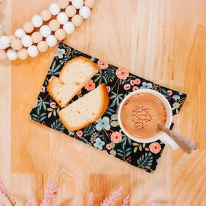 May include: A black floral fabric napkin with two slices of bread and a cup of hot chocolate on a wooden surface. A pink flower arrangement is in the lower left corner of the image.