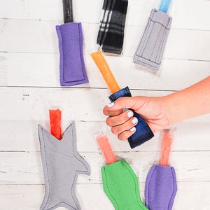 May include: A set of six colorful fabric pouches for holding popsicles. The pouches are made of felt and have different shapes, including a shark, a fish, and a rectangle. The pouches are filled with popsicles in various colors, including orange, red, and blue.