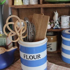 May include: A blue and white striped ceramic flour canister with the word "FLOUR" in black. The canister holds kitchen tools, including a woven whisk. Other vintage kitchen items are on shelves in the background.
