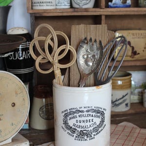 May include: A white ceramic jar with the text "James Keiller & Sons Dundee Marmalade" holds kitchen utensils, including a whisk and a decorative woven item. Other vintage jars and containers are on a wooden shelf.