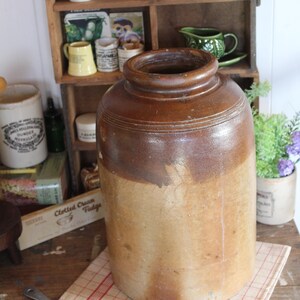 May include: An antique stoneware crock with a cylindrical body and a dark brown rim. The crock has a two-tone colour scheme, with a lighter tan base and a darker brown upper portion. A metal whisk rests on a red and white checked cloth.