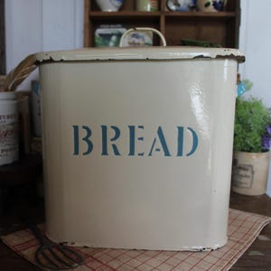 May include: Cream-colored metal bread box with a hinged lid and a handle. The word "BREAD" is stenciled in blue on the front. The bread box sits on a checkered cloth, with other vintage kitchen items in the background.