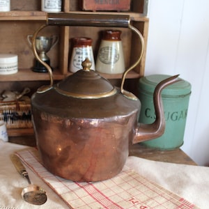 Puede incluir: Una tetera de cobre con asa y boquilla de latón se encuentra sobre un paño de lino con un estampado de cuadros rojos y blancos. La tetera forma parte de una colección de utensilios de cocina vintage.