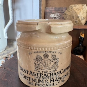 Könnte beinhalten: Ein beigefarbener Keramiktopf mit dickem Rand und einer kleinen Kerbe. Die Vorderseite zeigt ein schwarzes königliches Wappen mit Löwe und Einhorn sowie den Text BY APPOINTMENT TO H.M. THE KING FINEST ASTRACHAN CAVIAR IMPORTED BY FORTNUM & MASON 182 PICCADILLY.W.