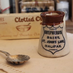 May include: A vintage cream pitcher with a brown rim and the text "SHEPHERD'S DAIRY, ST. JOHN'S LANE, HALIFAX" in black. A silver spoon rests on a white cloth, with a box of "Clotted Cream" in the background.