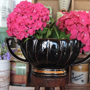 Puede incluir: Una jardinera de cerámica negra con ribetes dorados, llena de vibrantes flores de hortensia rosa. La jardinera tiene dos asas y se apoya en un pequeño taburete de madera. Otros objetos vintage, incluyendo tarros y libros, están en el fondo.
