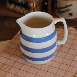 May include: A white ceramic jug with blue horizontal stripes. The jug has a handle and a spout. It is placed on a red and white checked cloth.