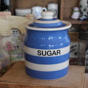May include: Blue and white striped ceramic sugar jar with a lid. The jar has the word "SUGAR" in black letters on the front. The jar is sitting on a wooden surface. Other kitchen items are in the background.