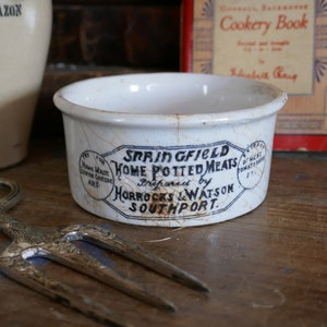 May include: A small, aged white ceramic bowl with a blue and black label reading "SPRINGFIELD HOME POTTED MEATS". A silver serving fork and a cookbook are in the background. The bowl shows signs of age and cracking.