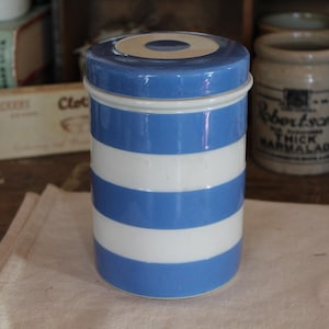 May include: A blue and white striped ceramic jar with a lid. The jar is cylindrical and has alternating bands of blue and white. The lid is blue with a white rim and a small circular indentation. The jar is sitting on a beige cloth.