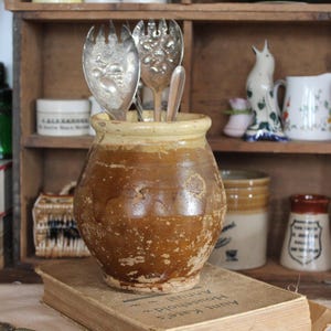 Op de afbeelding: Een bruine keramische pot met een lichtgele rand, met zilveren serveerbestek. De pot staat op een antiek boek. Andere vintage items zijn zichtbaar op de achtergrond, waaronder een houten plank.