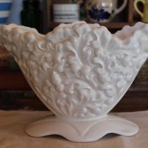 May include: A white ceramic bowl with a scalloped rim and intricate floral design. The bowl is made of a smooth, glossy material and has a wide, open mouth.