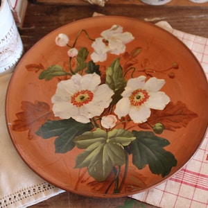May include: A round, orange-toned decorative plate featuring a hand-painted floral design. The flowers are white with orange centers, surrounded by green leaves and brown accents. A vintage serving spoon and utensil are visible.