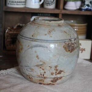Könnte beinhalten: Eine verwitterte, runde Keramikvase mit breiter Öffnung. Die Vase hat eine hellblau-graue Grundfarbe mit verblassten blauen Linien und braunen, rostähnlichen Flecken. Sie steht auf einem cremefarbenen Tuch, mit anderen Vintage-Artikeln im Hintergrund.