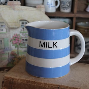 Peut inclure: Pichet à lait en céramique à rayures horizontales bleues et blanches. Le mot "MILK" est imprimé en noir sur le devant. Le pichet a une anse et un bec verseur. Il est posé sur un livre.