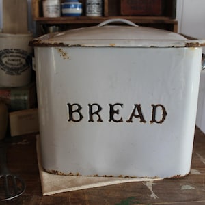 May include: A white rectangular bread box with a lid and handle. The word "BREAD" is stenciled in dark brown on the front. The box has a distressed, vintage appearance with rust-colored accents.