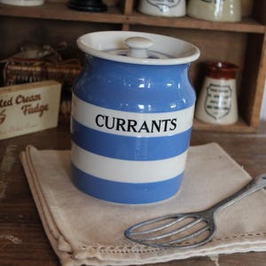 May include: A blue and white striped ceramic jar labeled "CURRANTS" with a lid, sitting on a beige linen napkin. A vintage metal serving spoon rests beside the jar. The jar is part of a collection of kitchenware.