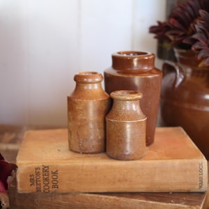 Puede incluir: Tres tarros cilíndricos de gres marrón de diferentes alturas con cuellos estrechos y bordes gruesos. Los tarros están dispuestos en grupo sobre un libro rectangular titulado Mrs. Beeton's Cookery Book, que tiene una cubierta marrón claro desgastada.