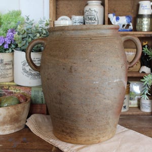 May include: A large, antique, brown stoneware jar with two handles. The rustic vessel is displayed on a wooden surface with other vintage items, including a small bowl, potted plants, and decorative jars. The jar has a textured surface.