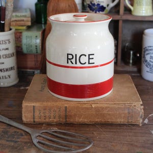 Peut inclure: Pot en céramique blanche pour le riz avec une bande rouge et le mot "RICE" imprimé en noir. Le pot a un couvercle et est posé sur un livre brun.