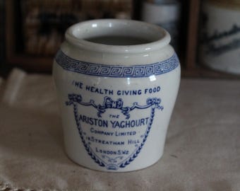 May include: A white ceramic jar with blue Greek key border and text. The jar reads "THE HEALTH GIVING FOOD" and "THE ARISTON YAGHOURT COMPANY LIMITED 18 STREATHAM HILL LONDON SW2" in blue script. The jar is on a white cloth.