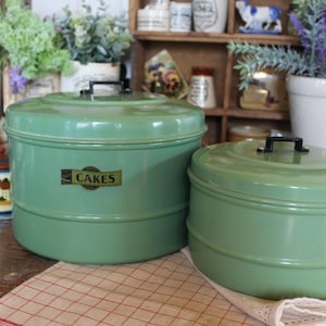 Peut inclure: Deux boîtes à gâteaux rondes en métal vert vintage avec des poignées noires. La plus grande boîte porte une étiquette « CAKES ». Elles sont de conception classique, parfaites pour conserver les pâtisseries. Elles sont posées sur une surface en bois.