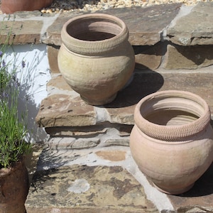 Peut inclure: Deux pots en terre cuite beige de forme arrondie avec une large ouverture et un bord texturé et nervuré. Les pots présentent un corps bombé et une finition mate et terreuse. Un troisième pot partiellement visible avec de la lavande est à gauche.
