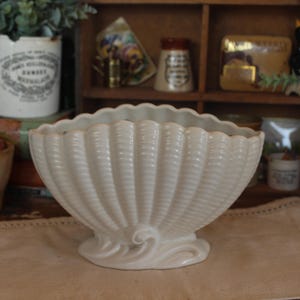 May include: A cream-colored, seashell-shaped ceramic bowl with a scalloped rim and textured vertical lines. The bowl sits on a decorative base with a swirling design. The background includes other vintage items on a wooden shelf.