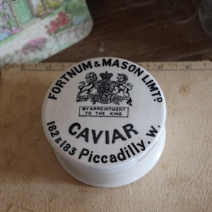Puede incluir: Un tarro de caviar de cerámica blanca con texto negro y un escudo real. El tarro lleva la inscripción "FORTNUM & MASON LIMTD" y "CAVIAR Piccadilly, w." El tarro está sobre una superficie marrón claro.