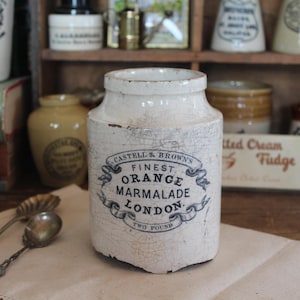 May include: Antique white ceramic jar with a distressed finish, featuring a blue and white label that reads "Finest Orange Marmalade London." The jar is labeled "Two Pound" and is part of a collection of vintage kitchenware.