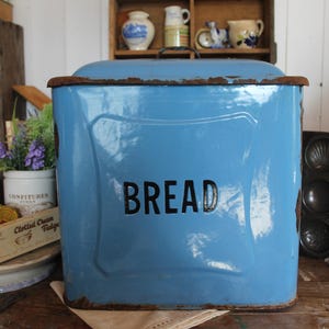Peut inclure: Une boîte à pain bleu clair avec le mot "BREAD" en noir. Le récipient rectangulaire a un couvercle incurvé et présente des signes d'usure. D'autres ustensiles de cuisine vintage sont visibles en arrière-plan.