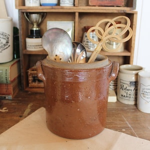 Puede incluir: Un tarro de cerámica marrón con asas, que contiene utensilios de cocina. El tarro está sobre una superficie de madera, con otros artículos de cocina vintage en el fondo, incluyendo tarros y un trofeo.
