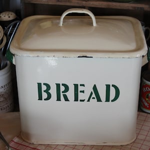 May include: Cream-colored bread box with a matching lid and a handle. The word "BREAD" is stenciled in green on the front. The box is rectangular and appears to be made of metal.