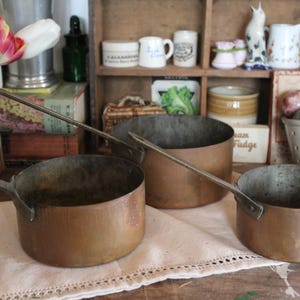 Peut inclure: Trois casseroles anciennes en cuivre avec de longues poignées reposent sur un tissu crème. Les casseroles ont une patine chaude et vieillie. Une étagère à l'arrière-plan présente des ustensiles de cuisine vintage, dont des pots, des tasses et un petit pichet en céramique.
