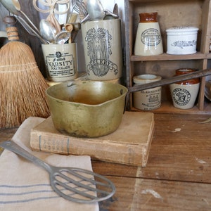 May include: A vintage brass pot with a long handle sits on a stack of books. A small whisk sits on a white and blue striped towel. The pot is surrounded by antique jars and a broom.