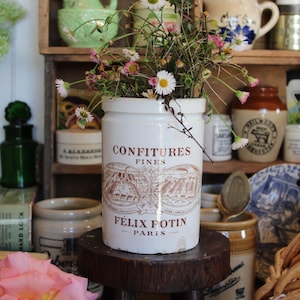May include: A white ceramic jar with the text "CONFITURES FINES FÉLIX POTIN PARIS" and a landscape illustration, filled with wildflowers. Other vintage jars and containers are in the background, creating a rustic, antique aesthetic.
