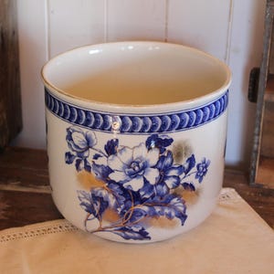 Puede incluir: Una maceta cilíndrica de cerámica con base color crema que presenta un patrón floral azul y un borde decorativo azul en la parte superior. El diseño incluye flores y hojas azules con sutiles acentos dorados sobre la superficie blanca.