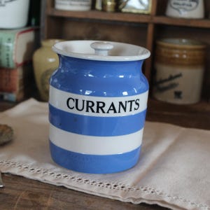 May include: A vintage ceramic jar with a blue and white striped design, labeled "CURRANTS" in black lettering. The jar has a white lid and sits on a white cloth. A small silver spoon is in front of the jar.