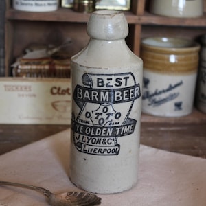 May include: A vintage, cream-coloured ceramic bottle with a cylindrical form and a narrow neck. The bottle has a black and white label that reads "BEST BARM BEER" and "YE OLDEN TIME". The bottle is displayed on a light-coloured cloth.