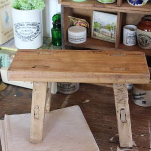 Puede incluir: Un pequeño taburete de madera rústico con una parte superior rectangular y cuatro patas. El taburete está hecho de madera de color claro con vetas visibles y mide aproximadamente 30 cm de largo. Tiene un diseño simple y artesanal, perfecto para una estética vintage o de granja.