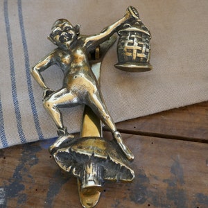 May include: A brass door knocker in the shape of a mythical creature holding a lantern. The creature is standing on a mushroom-shaped base.