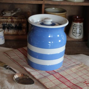 May include: A blue and white striped ceramic jar with a lid. The jar is sitting on a red and white checkered cloth, with a silver spoon in the foreground. Other vintage kitchen items are in the background.