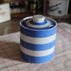 May include: A blue and white striped ceramic sugar bowl with a lid and a small spoon slot. The bowl is cylindrical and sits on a red and white checkered cloth. The lid has a small, round knob.