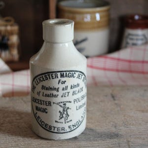 May include: A small, off-white ceramic bottle with a black and white label. The label reads "LEICESTER MAGIC JET ST" and "For Staining all kinds of Leather JET BLACK POLISH." The bottle is likely an antique, with a vintage aesthetic.