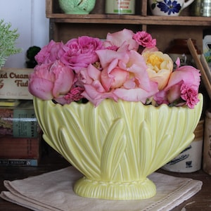 Pode incluir: Um vaso amarelo pálido, em estilo art déco, cheio de rosas cor-de-rosa e amarelas. O vaso tem um design texturizado em forma de leque. As flores estão em plena floração, criando uma exibição vibrante. O vaso está sobre um pano branco.