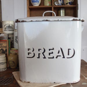 Puede incluir: Panera de esmalte blanco con tapa y asa. La palabra "BREAD" está recortada en la parte delantera. La panera tiene forma rectangular y está sobre una superficie de madera. Parte de una colección de utensilios de cocina vintage.