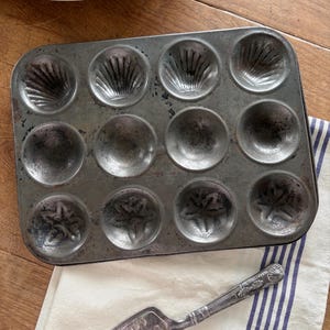 May include: A rectangular metal baking pan with twelve molded cavities featuring shell and floral patterns, accompanied by a silver-toned metal cake server with an ornate handle and an engraved blade design, both showing signs of age and wear.